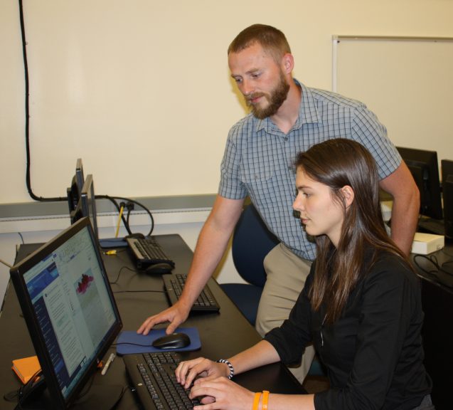 Kytt MacManus, standing left, and Dorothee Grant, sitting right, look at a computer monitor as they study images of nightime lights data.